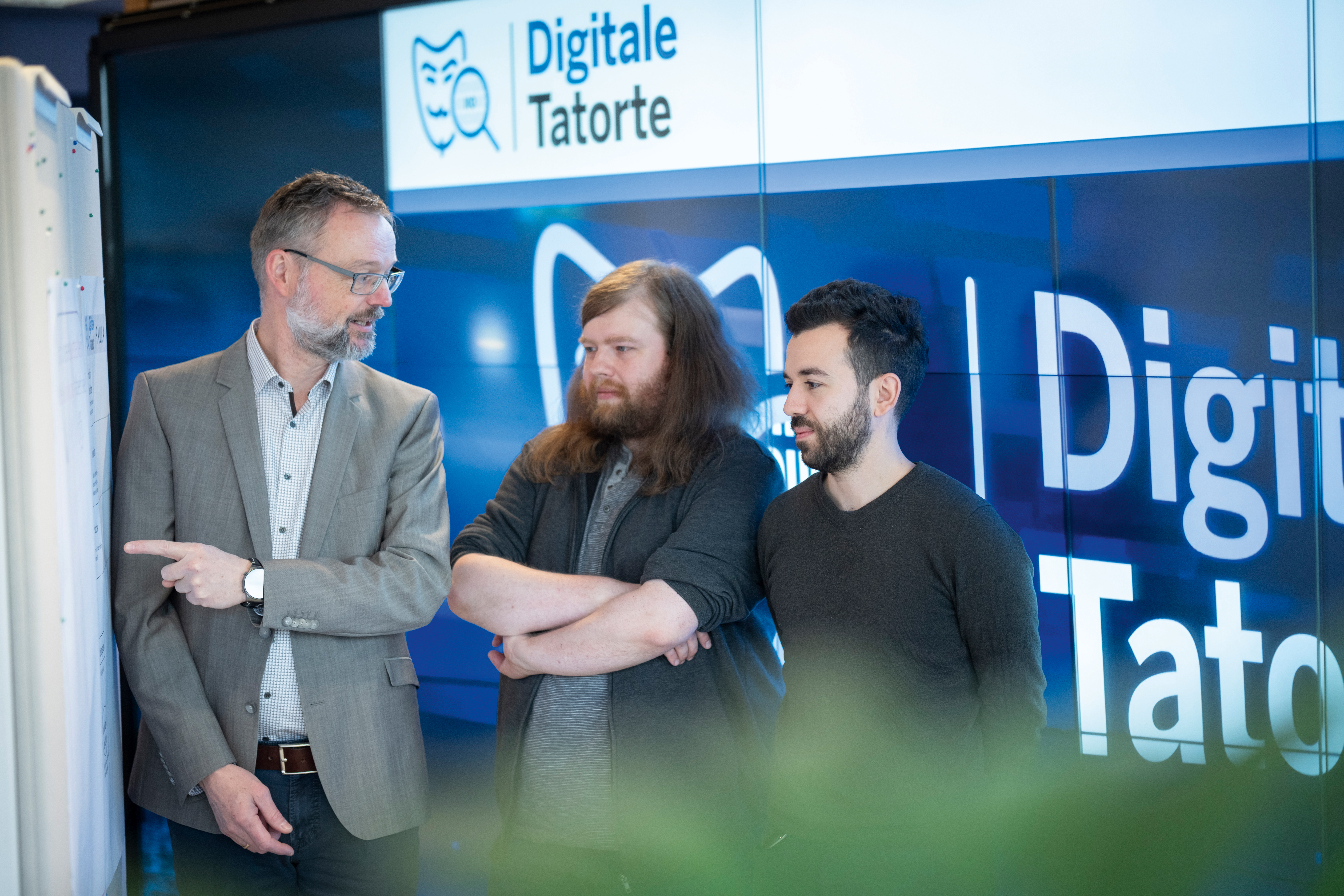 Andreas Bruns, Leiter des Projektbüros „Digitale Tatorte“, erläutert Robin Marchel und Burak Uslu aus dem Tatort-Team Dortmund die Aufstellung der neuen Einheit. Im Hintergrund ist eine Bildschirm-Wand mit einer blau-weißen Grafik mit der Aufschrift "Digitale Tatorte" zu sehen.