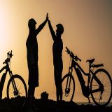 Die Silhoutte zweier E-Bike-Fahrer - ein Mann und eine Frau - und ihrer Fahrräder vor dem Hintergrund einer untergehenden Sonne. Es wirkt so, als hätten sie gerade euphorisch einen Berg erklommen.
