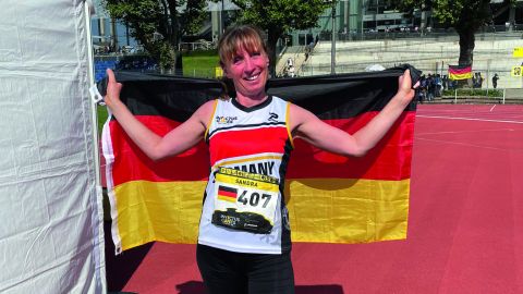 Die Polizistin Sandra Winkler steht im Deutschland-Trikot auf einem Sportplatz. Über ihren Rücken hält sie eine Deutschlandflagge. Im Hintergrund sind Bäume und ein Ausschnitt der Düsseldorfer Arena zu sehen