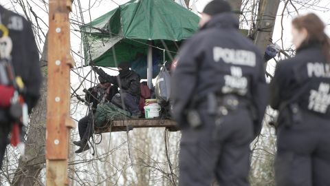 Im Hintergrund ist eine Plattform zu sehen, die Aktivisten zwischen zwischen Bäumen errichtet haben. Auf der Plattform befinden sich mehrere Aktivisten. Im Vordergrund sind verschwommen Polizistinnen und Polizisten von hinten zu sehen.