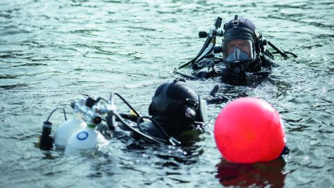 Zwei Taucher und ein Polizeiboot im Wasser, ein Fundort wird mit einer Boje markiert.