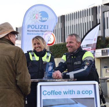 Eine Poliizeibeamtin und ein -beamter im Gespräch mit einem Mann mit Hut. Vorne ein Plakat mit den Worten "Coffee with a Cop", im Hintergrund ein Aufsteller der Polizei MK.