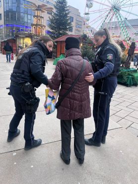 Zwei Polizeibeamtinnen sprechen mit einer Seniorin, die nur von hinten zu sehen ist, am Rand eines Weihnachtsmarktes.