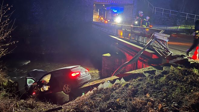 Am Fuße einer Brücke liegt ein Pkw nach vorn gekippt im Wasser eines Flusses. Auf der Brücke stehen Rettungskräfte. 